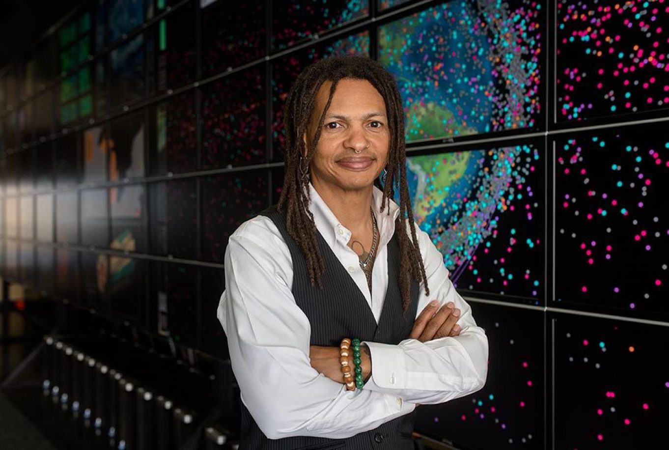 Moriba Jah stands with arms folded in front of an image of outer space.